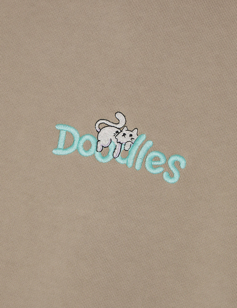 Sand-colored Doodles Mello Hoodie featuring an embroidered design of Mello lounging atop the Doodles logo, made from heavyweight 100% cotton for comfort and style. Logo close-up.